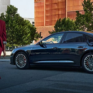A black Audi is parked on a street, partially plugged into a charging station. Trees and a modern building are visible in the background.