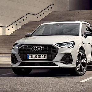 A grey Audi SUV is parked at a charging station beside a modern building. The scene suggests innovation and sustainability against a scenic backdrop.