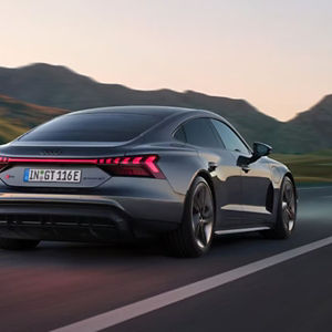 A grey Audi car driving on a road, with mountains in the background at sunset. The car's design conveys speed and elegance.