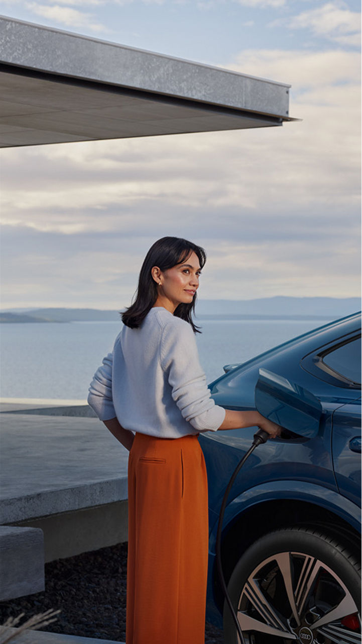 Woman in white sweater and orange skirt charges blue electric car by the sea. She smiles slightly, conveying a sense of sustainability and calm.