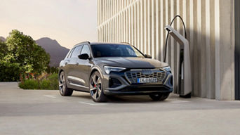 A grey electric Audi vehicle plugged into a charging station outside a modern building