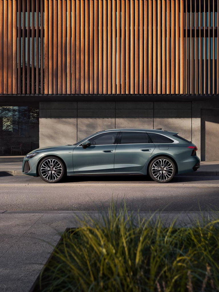Auf dem Bild ist ein Audi A6 Avant e-hybrid zu sehen. Das Äußere des Fahrzeugs ist in einer einzigartigen Metallic-Farbe gehalten, die zur Umgebung passt. Der Hintergrund zeigt ein modernes Gebäude mit einer Betonwand und Holzelementen.