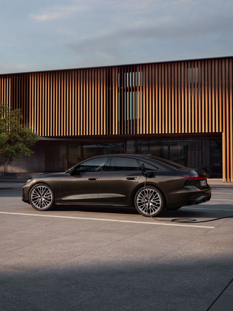 Das Bild zeigt eine braune (eine Audi exclusive Farbe) Audi A6 Limousine e-hybrid, der an eine Stromtankstelle angeschlossen ist. Im Hintergrunde ist moderne Architektur zu sehen. 