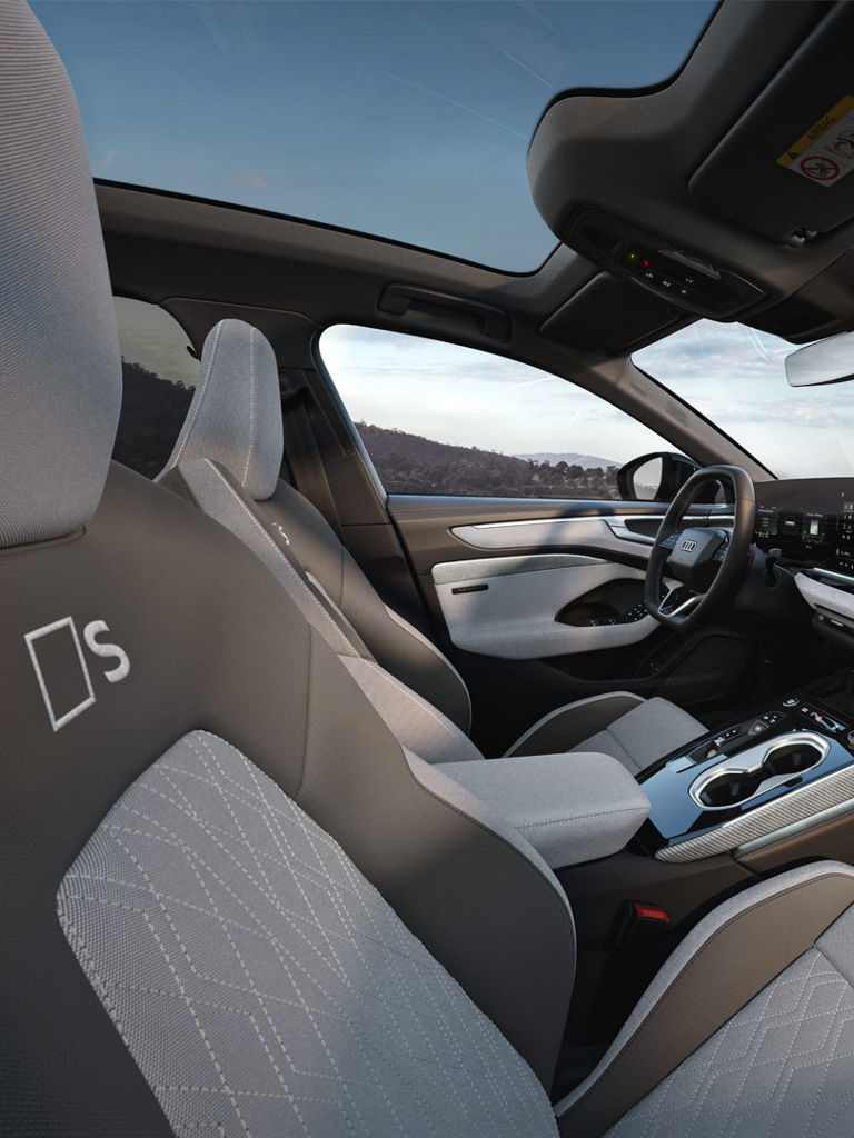 Das Bild zeigt den Innenraum einer Audi A6 Limousine e-hybrid. Der Innenraum ist zweifarbig gestaltet, hauptsächlich in Grau und Weiß. Die Mittelkonsole wird von einem großen Touchscreen-Display dominiert. Die Sitze sind mit einem „S“-Logo gekennzeichnet, was auf eine sportliche Ausstattungsvariante hindeutet. Das Armaturenbrett zeigt ein integriertes Display für Navigations-, Medien- und Fahrzeuginformationen.