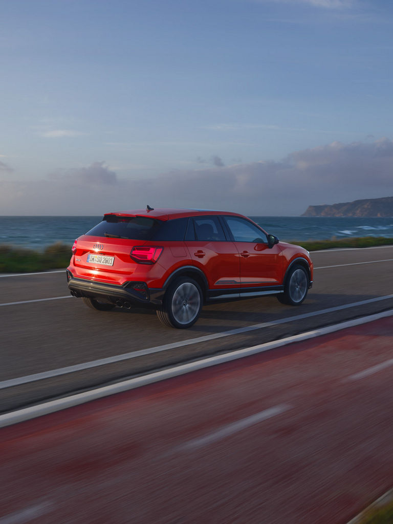 Ein roter Audi SQ2 fährt auf einer Küstenstraße am Meer entlang. Weiter hinten sieht man eine Klippe.
