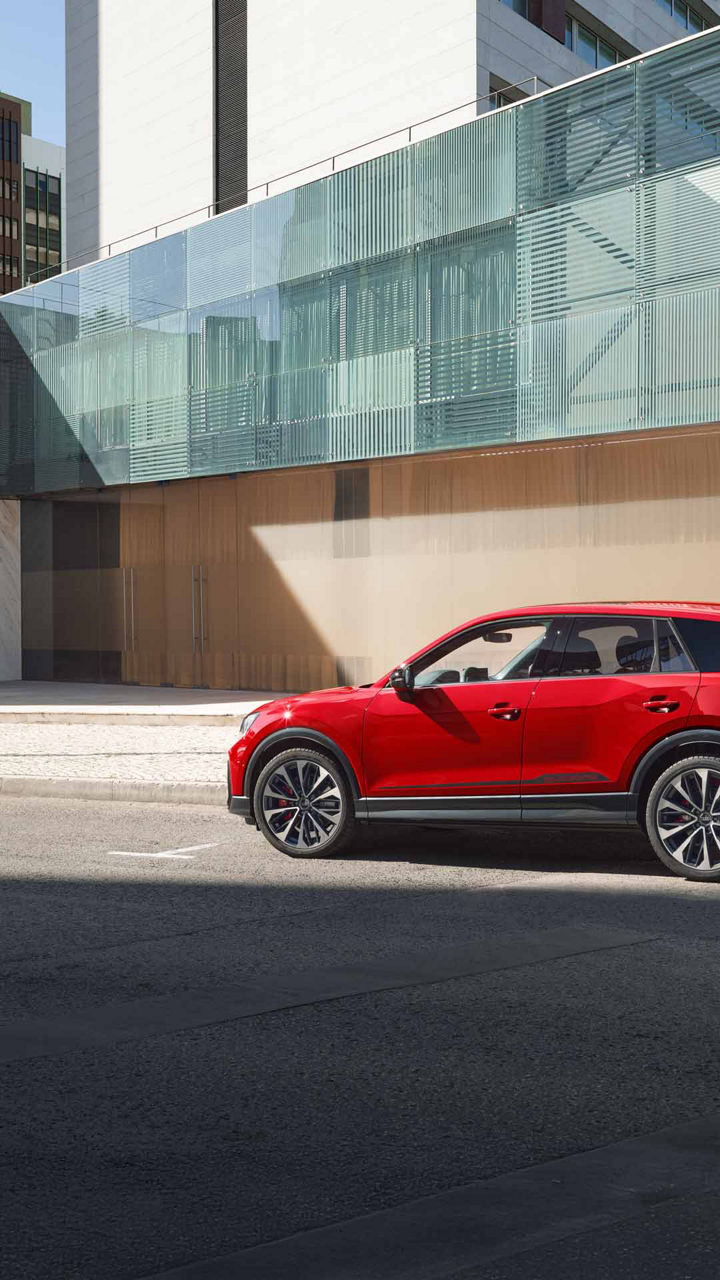 Ein roter Audi SQ2 seitlich auf städtischer Straße vor modernem Gebäude mit Glas- und Steinfassade geparkt.