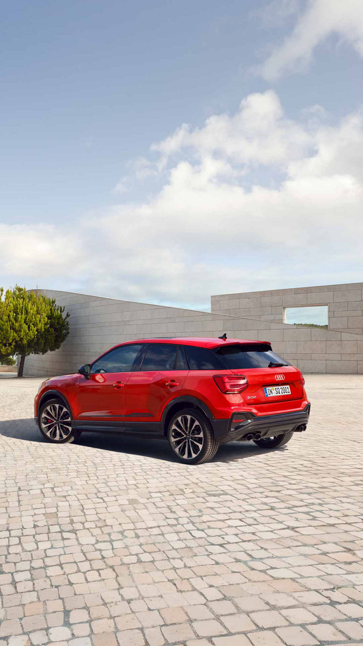 Ein roter Audi SQ2 von hinten auf Platz mit Bäumen, lächelnde, brünette Frau in grauem, langen Mantel läuft daran vorbei.