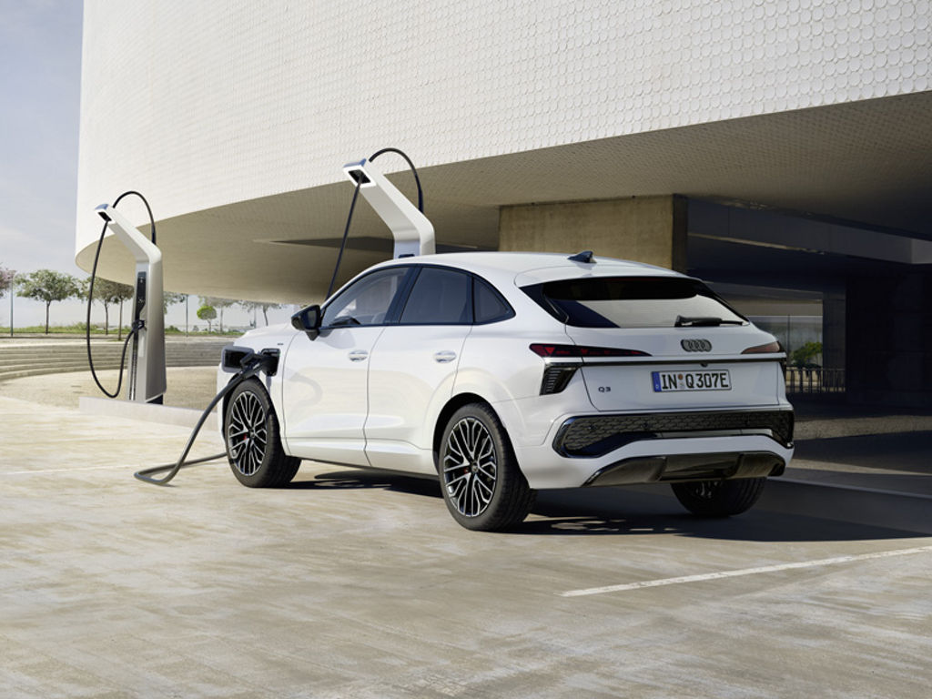 Ein weißer Q3 Sportback e-hybrid lädt Strom an einer Ladesäule vor einem modernem Gebäude, das Fahrzeug wirft lange Schatten.