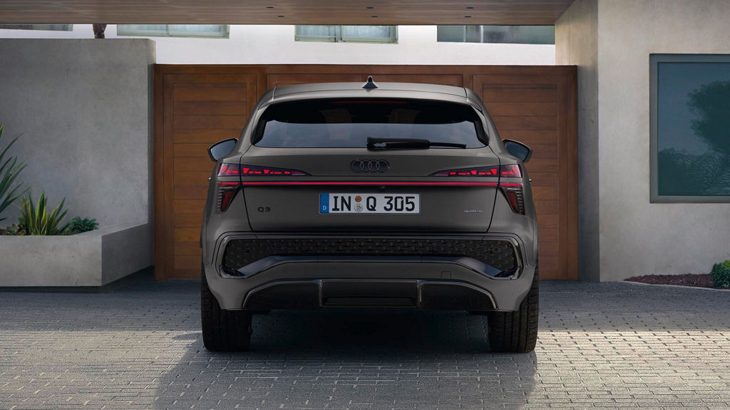 Der Audi Q3 Sportback ist in einer Heckansicht zu erkennen. Er steht auf einem Parkplatz vor einem Hölzernen Eingangstor zu einem Wohnhaus.