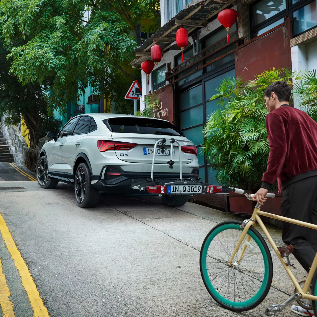 Ein weißer Audi Q3 Sportback mit Fahrradträger steht an steiler Straße, asiatischer Mann schiebt sein Fahrrad im Vordergrund.