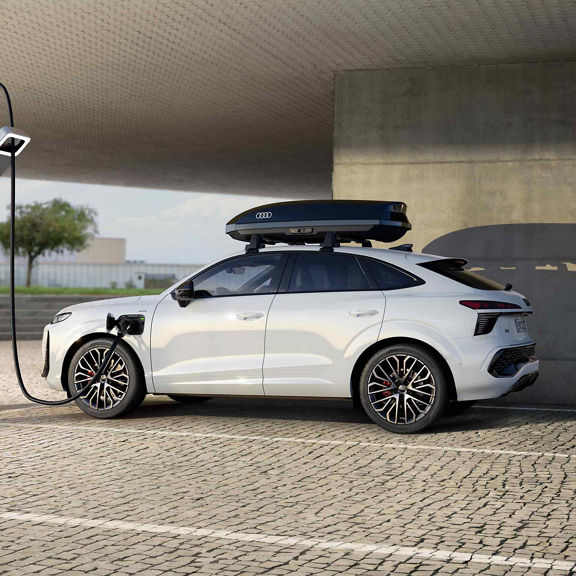 La Q3 Sportback e-hybrid, équipée d'un coffre de toit noir brillant, se recharge dans une station publique. Des bâtiments sont visibles en arrière-plan.