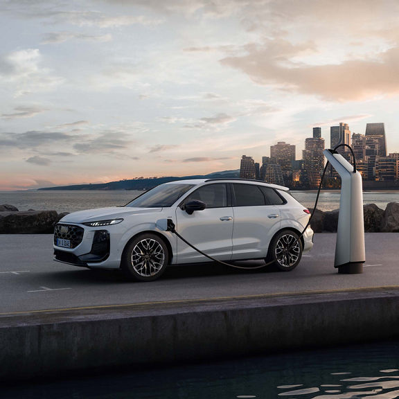 Audi Q3 SUV e-hybrid con due persone che scattano una foto con il loro cellulare. Si trova di lato di fronte all’acqua e si carica