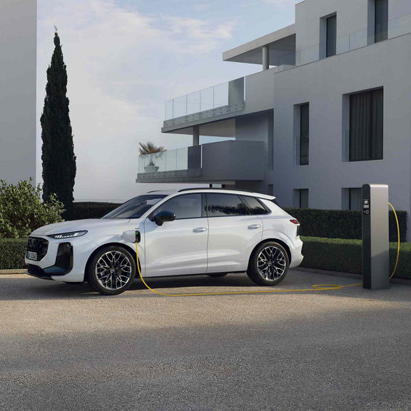 Sānskats uz baltu Q3 SUV e-hybrid, kas tiek lādēts stāvvietā pie modernas mājas.
