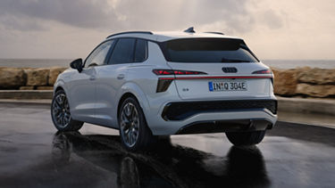 A white Q3 SUV e-hybrid drives along a wet road along the coast.	