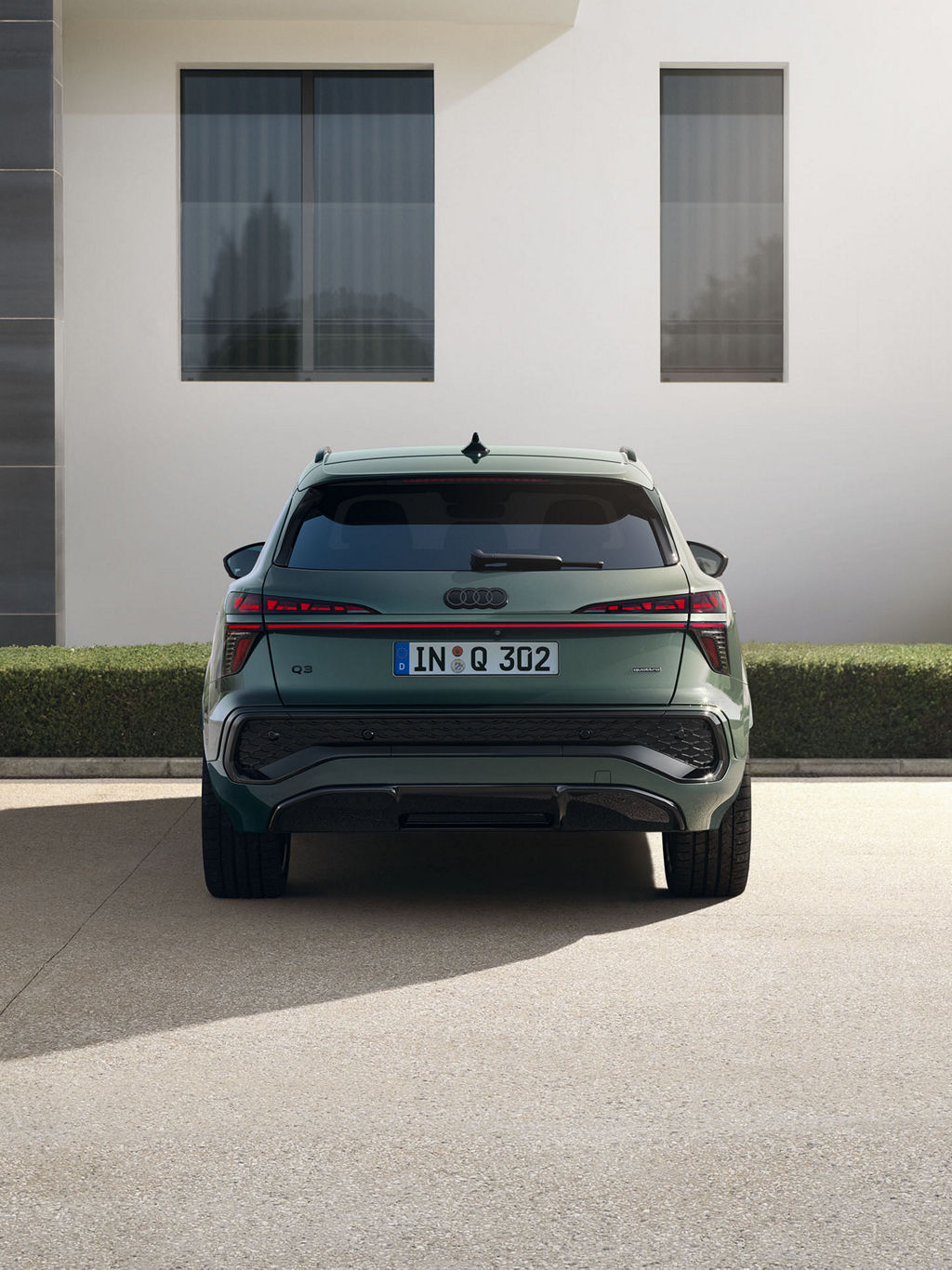 Das Bild zeigt einen grünen Audi Q3 SUV von hinten, vor einem modernen weißen Gebäude mit rechteckigen Fenstern und etwas Grün. Das Auto hat markante Rückleuchten und einen Heckstoßfänger.
