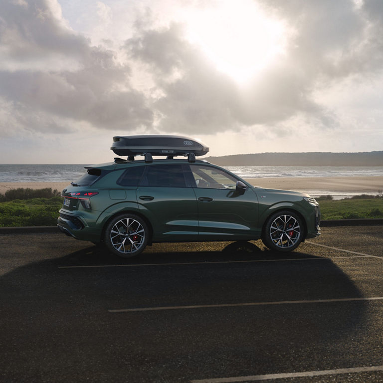 Das Bild zeigt einen grünen Audi Q3 SUV, der in der Nähe eines Strandes unter einem bewölkten Himmel geparkt ist. Auf dem Dachgepäckträger des Fahrzeugs ist eine Dachbox mit dem Audi-Logo montiert. Die Räder des Fahrzeugs haben sichtbare rote Bremssättel, was auf eine leistungsstarke oder sportliche Ausstattung hinweist.