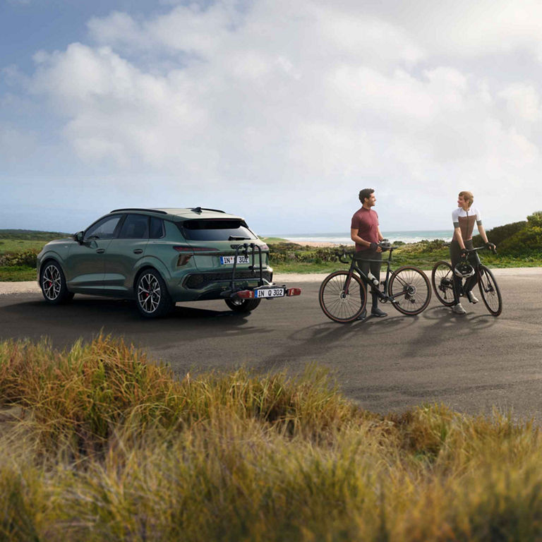 Das Bild zeigt einen grünen Audi Q3 SUV mit einem Fahrradträger am Heck, was darauf hindeutet, dass er für Outdoor-Aktivitäten ausgerüstet ist. Neben dem Auto ist ein Paar mit Fahrrädern zu sehen. Im Hintergrund ist eine Küstenlandschaft mit einem Strand zu sehen.
