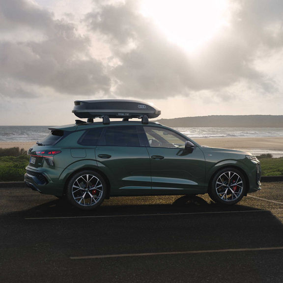 Seitliche Ansicht des Q3 SUV mit einer brillantschwarzen Dachbox, angestrahlt von der Sonne mit dem Meer im Hintergrund.