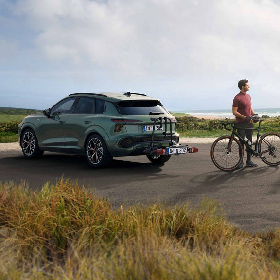 Der Q3 SUV mit Fahrradträger steht vor einer Küstenlandschaft, daneben ein junger Mann mit einem Gravelbike.