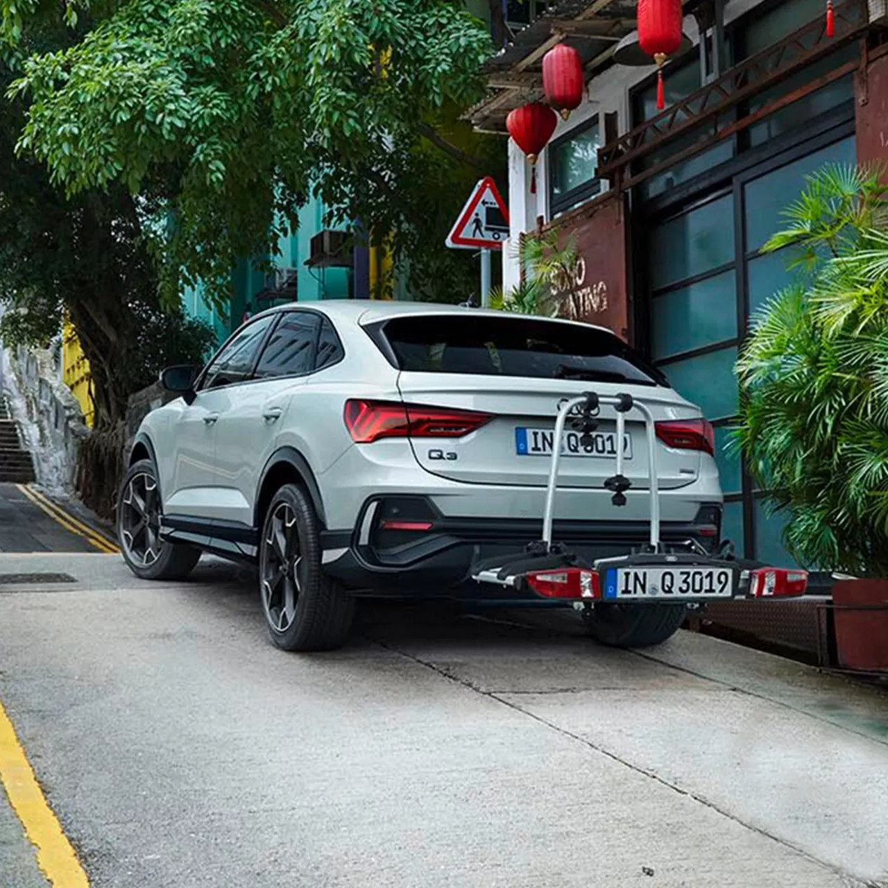 Ein weißer Audi Q3 Sportback mit Fahrradträger steht an steiler Straße, asiatischer Mann schiebt sein Fahrrad im Vordergrund.