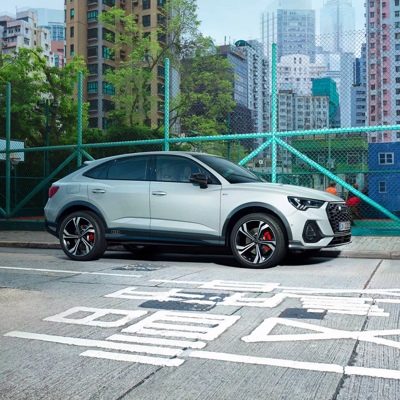 Ein weißer Audi Q3 Sportback vor hellgrünem Zaun, bunte Hochhäuser im Hintergrund.