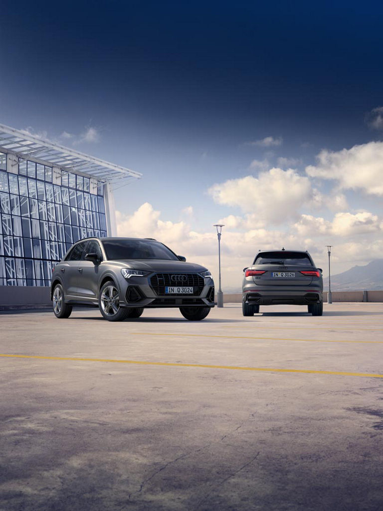 Zwei graue Audi Q3 stehen auf einem Dachparkplatz, einer von vorn, einer von hinten, modernes Gebäude im Hintergrund.