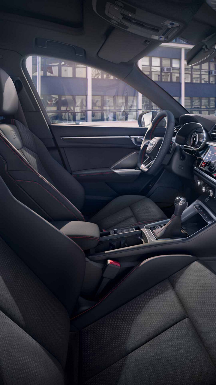 Blick auf Vordersitze und Cockpit des Audi Q3 mit schwarzer Innenausstattung und roter Ziernaht, Gebäude außen sichtbar.