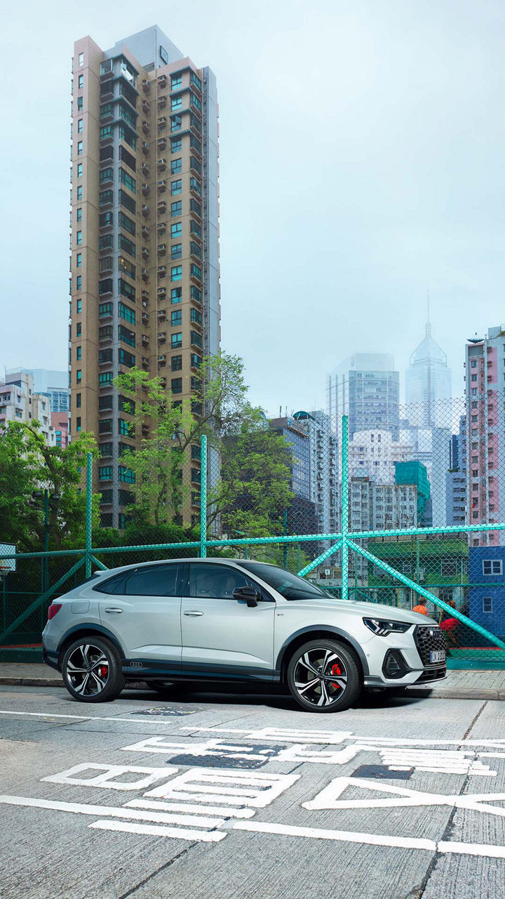 Ein weißer Audi Q3 Sportback vor hellgrünem Zaun, bunte Hochhäuser im Hintergrund.