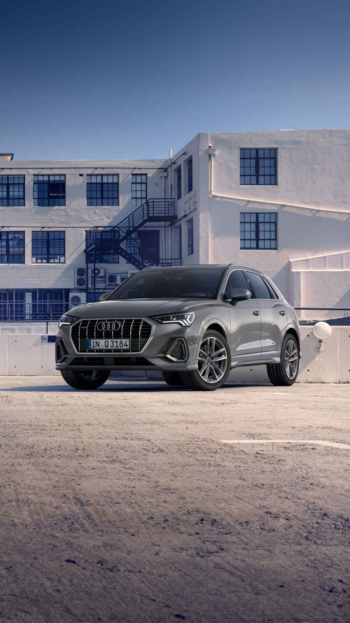 Ein grauer Audi Q3 steht vor weißem Industriegebäude mit dunkelblauen Fenstern und Außentreppen.