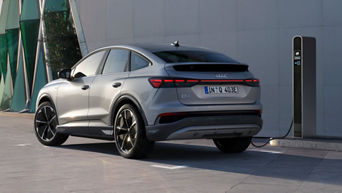 A silver Q4 Sportback e-tron with sport rims charges at a charging station in front of a modern building, the vehicle casting long shadows.