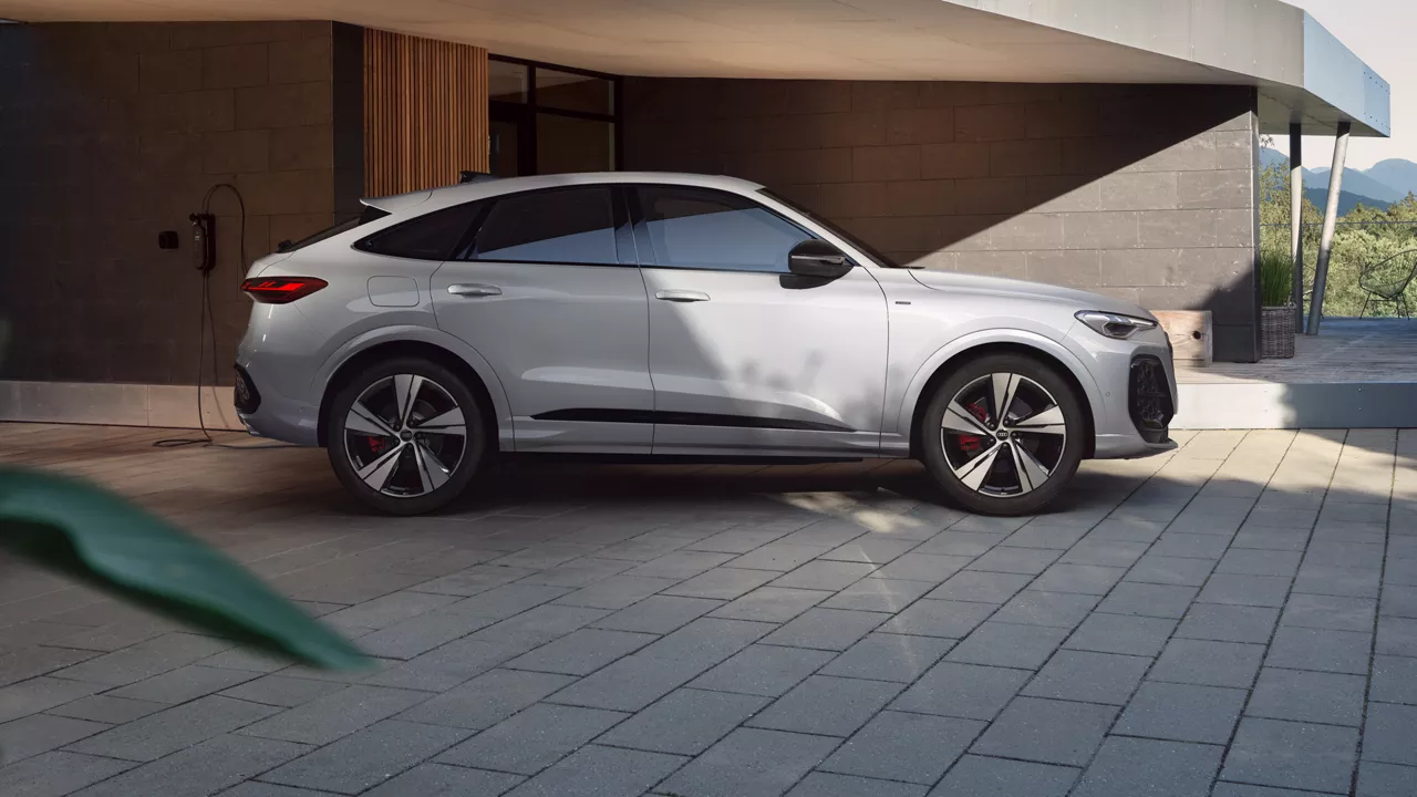 Weißer Audi SUV-Coupé lädt an einer Wandladestation vor einem modernen Wohnhaus mit Naturblick.
