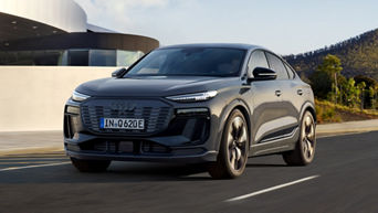 Der Q6 Sportback e-tron fährt auf einer Straße, im Hintergrund moderne Gebäude und ein Waldgebiet.