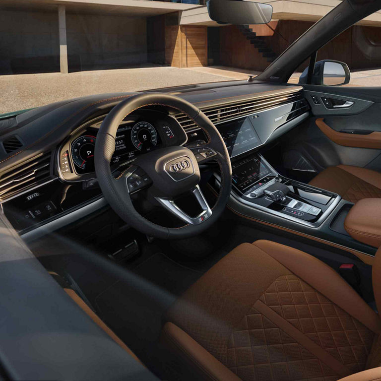 Innenraum eines Audi Q7 SUV mit braunen Leder, Sportsitzen, digitalem Cockpit und Lenkrad, Blick auf die Fahrerseite geparkt vor einem modernen Haus.
