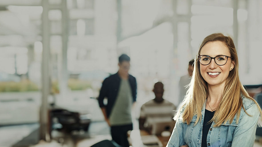 Young woman with blonde long hair, denim jacket and glasses stands with her arms crossed and smiles into the camera