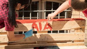 Two people paint AIA on a wooden pallet with brushes in red paint