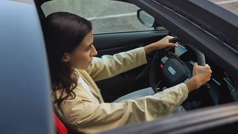 Dark-haired woman with beige jacket sits at the wheel of an Audi model, bird's eye view