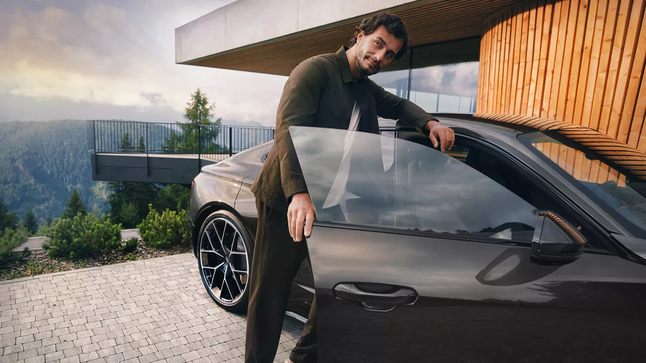 A dark-haired man in a suit gets into a dark Audi S e-tron GT on the passenger side, with a landscape of forests in the background