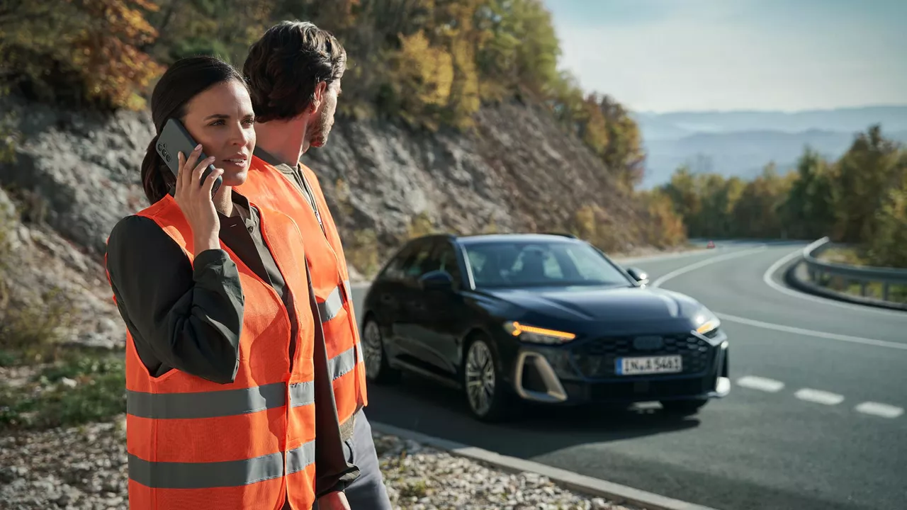 Eine Frau und ein Mann, die beide eine Warnweste tragen, stehen neben einer Straße. Der Mann blickt nach hinten auf einen Audi, dessen Warnblinkanlage eingeschaltet ist, während die Frau mit einem Handy in ihrer Hand telefoniert.