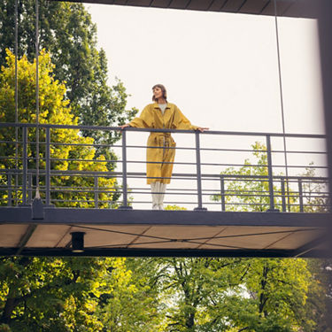 Woman in a yellow coat stands on a bridge, holding on to the railing and looking out into nature. In the background are trees with green leaves