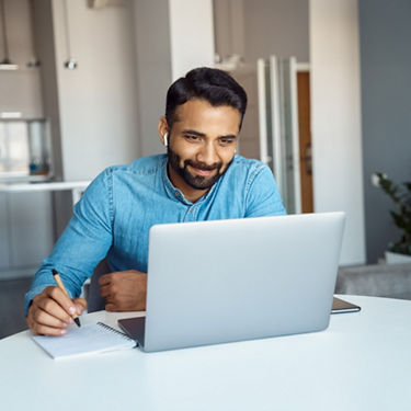 Young man with dark hair and a dark beard sits in front of a laptop and writes something on a piece of paper