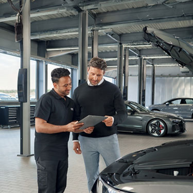A service employee stands with a customer in a workshop and shows the customer a document. In the background are Audi models