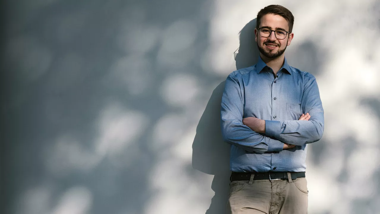 Marco steht mit verschränkten Armen vor einer Wand und lächelt in die Kamera