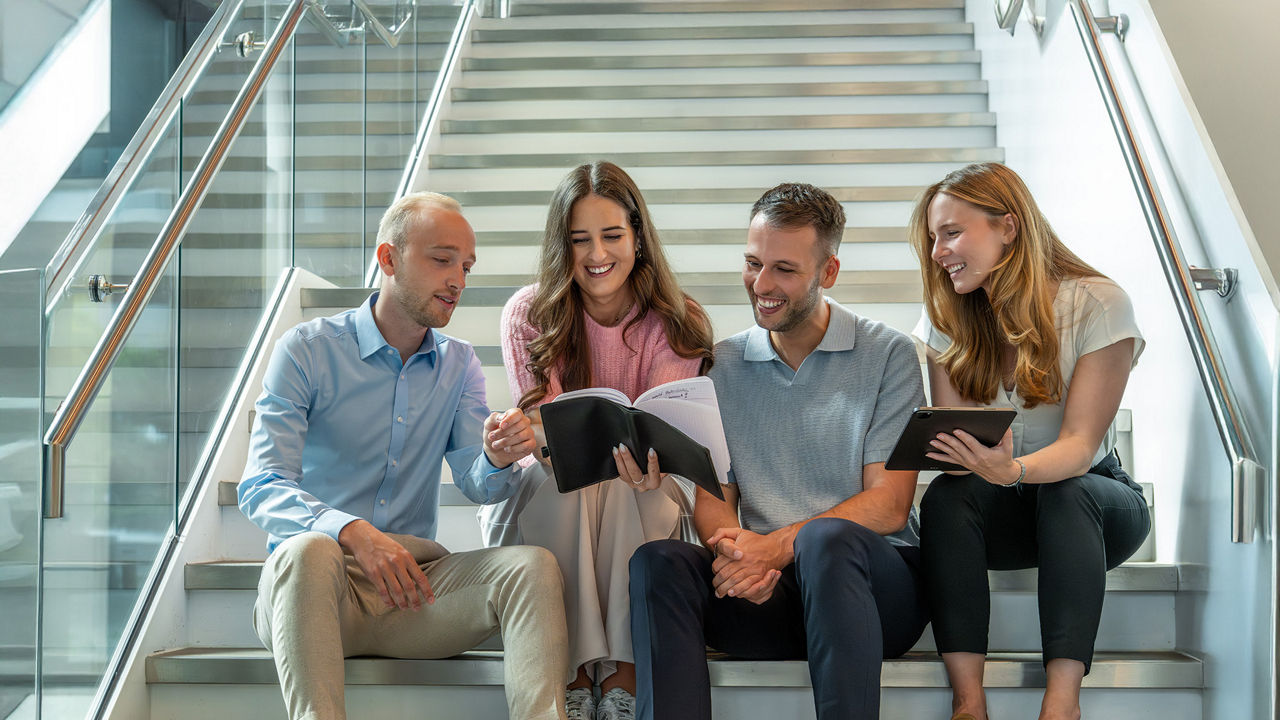 Four graduates are sitting on stairs and look into a book that one graduate is holding up