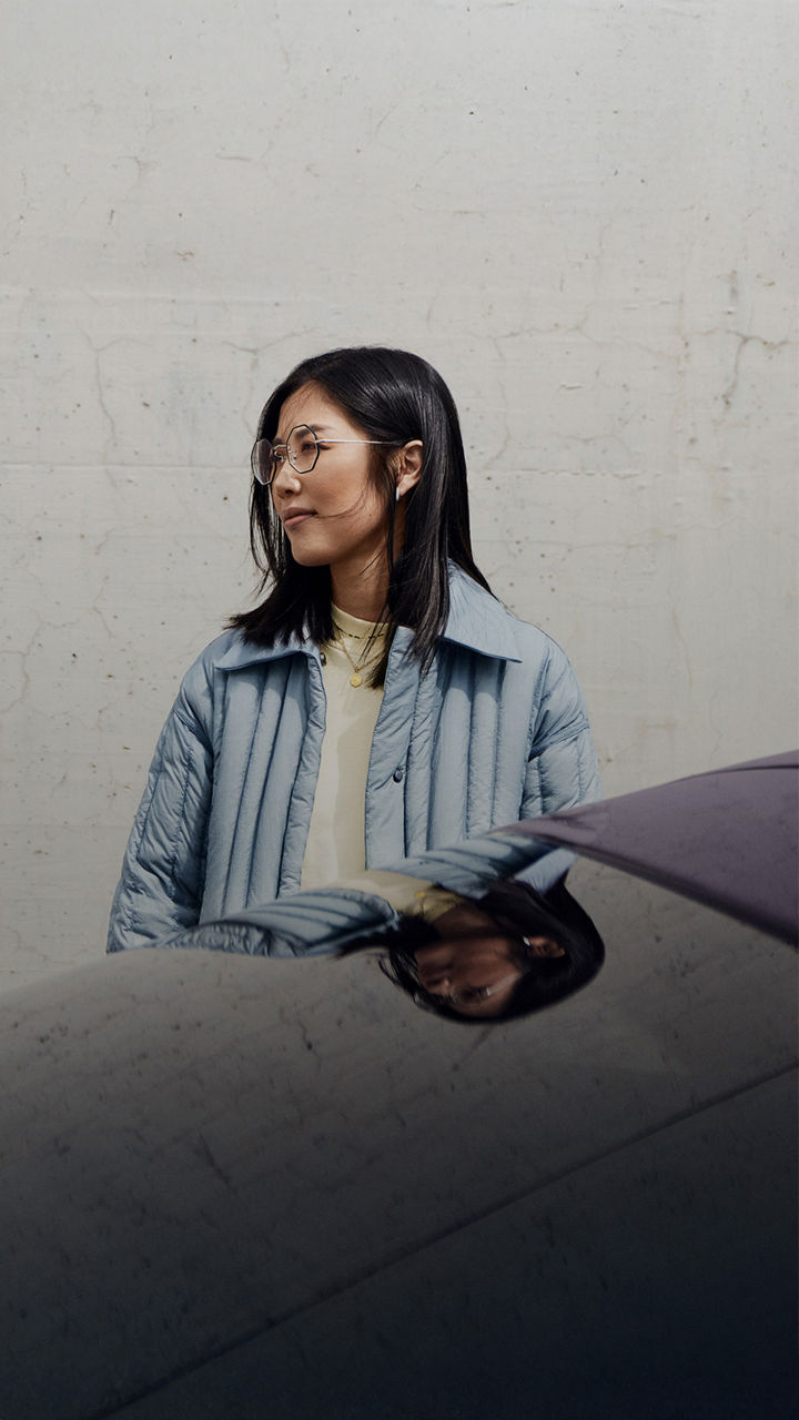 A woman in a blue jacket stands behind a sporty vehicle rear in front of a grey concrete wall and looks to the side. 