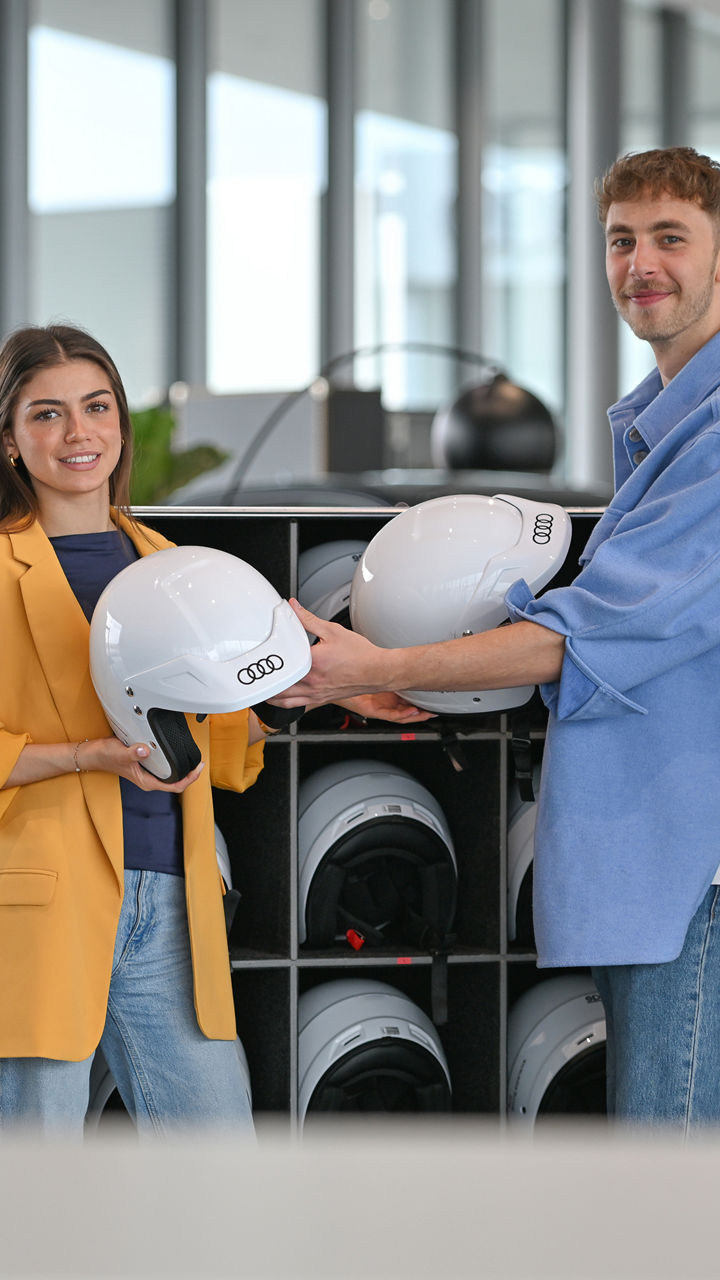 Zwei Auszubildende reichen sich weiße Helme mit einem Audi Logo