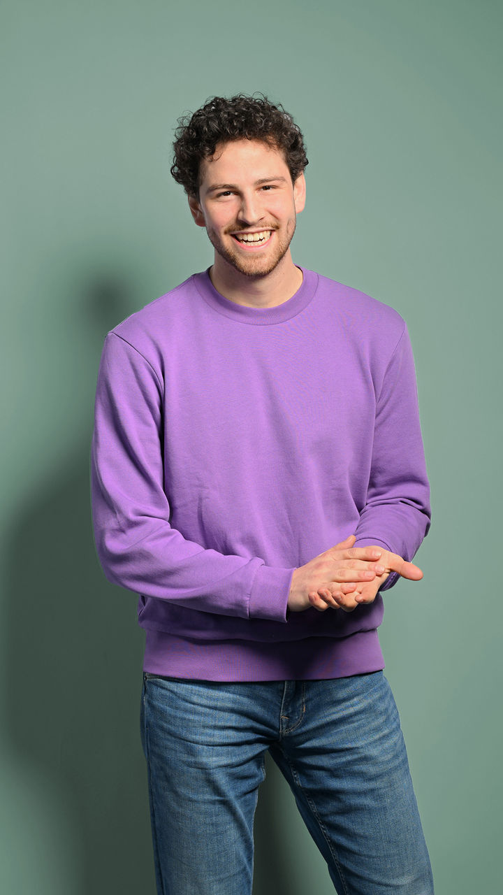 Fabian steht mit einem lila Pullover vor einer Wand und lacht an der Kamera vorbei