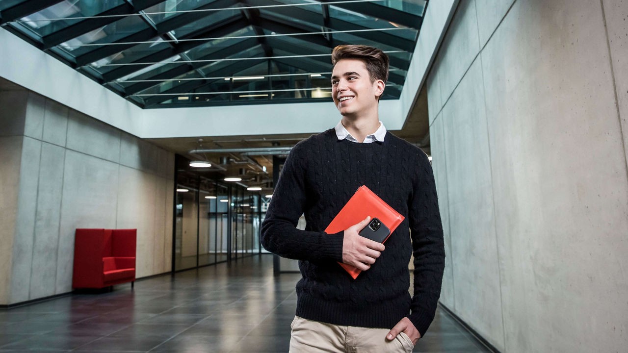 Ein Junge hat ein rotes Buch und ein Handy in der Hand und schaut lächeln an der Kamera vorbei