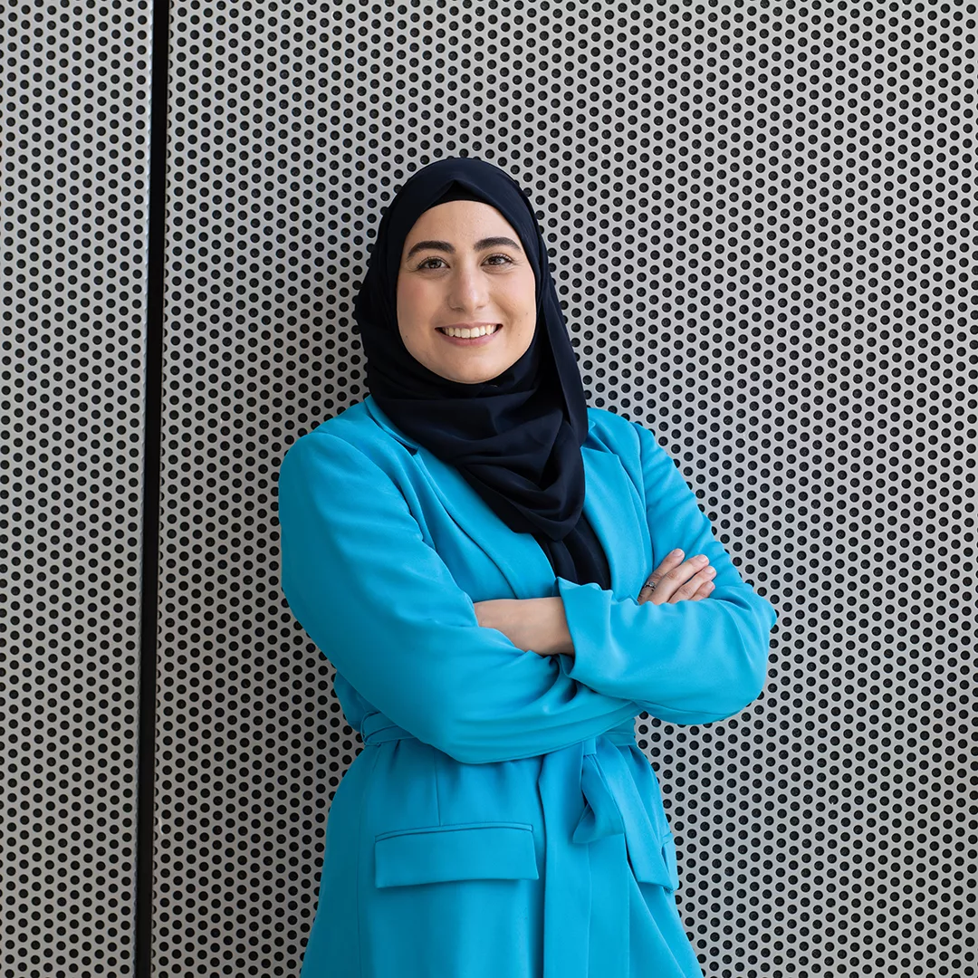 Betül stands smiling with her arms crossed in front of a wall