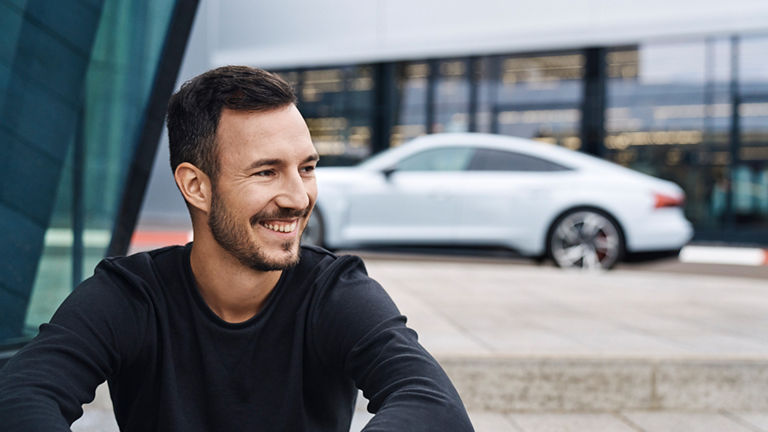 Andreas schaut lachend nach rechts und hat einen weißen Audi im Hintergrund