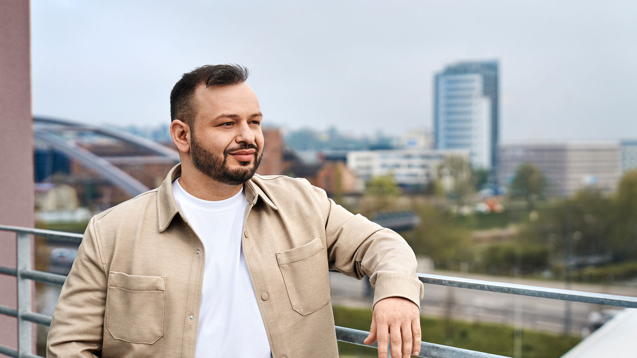 Buenyamin looks to the right and is leaning against a railing. He has a view of a city behind him